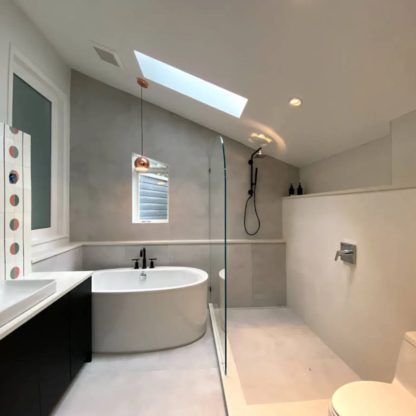 The same ensuite after renovation, with a freestanding tub, walk-in glass shower, large-format grey stone, and a copper pendant